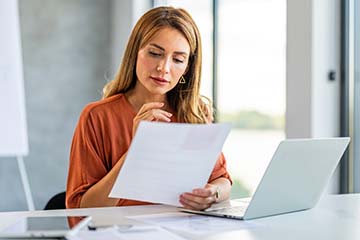 A woman looks at paper work while sitting in front of her laptop