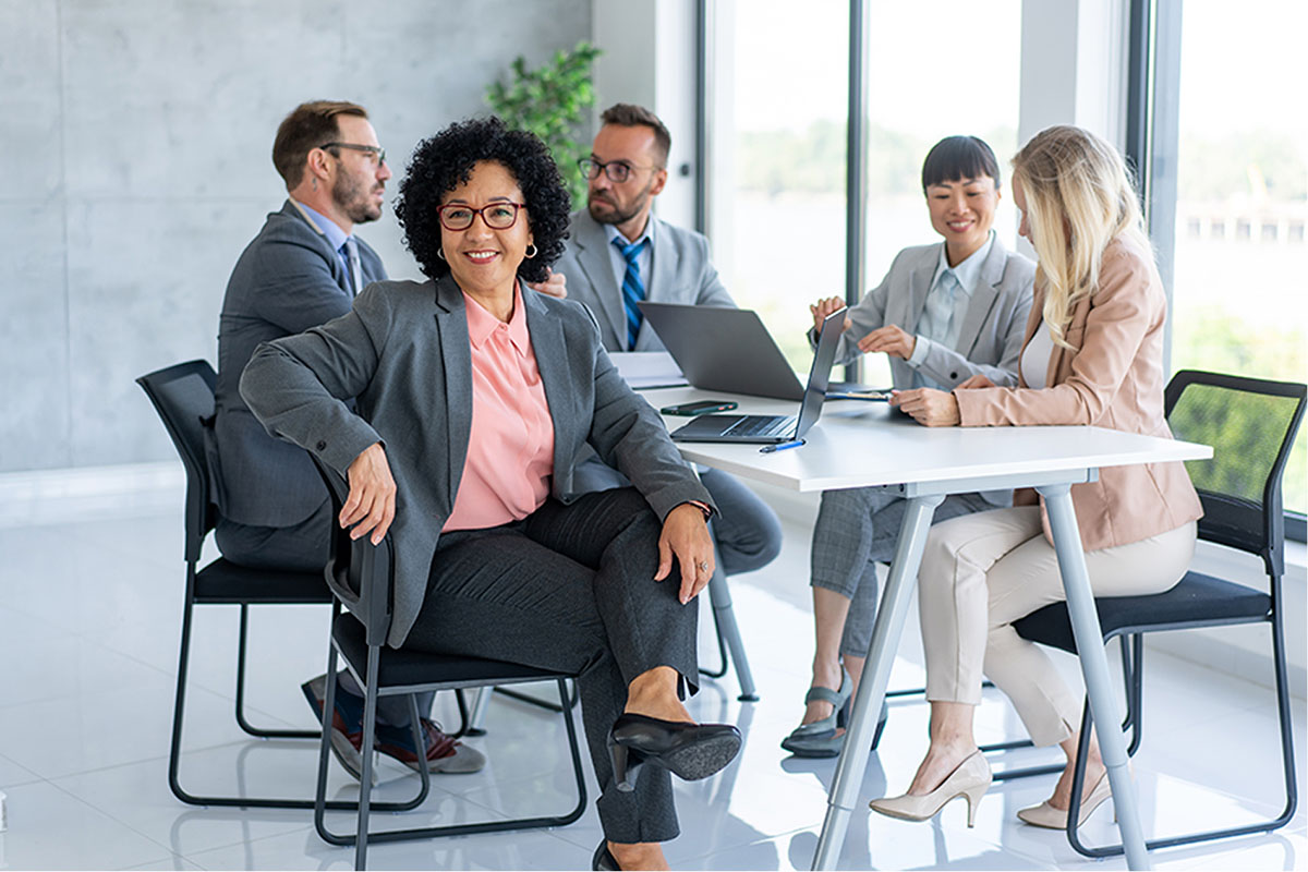 A group of business professionals sit a table, a woman with glasses looks into the camera